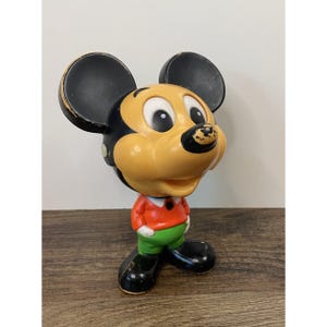 May include: A vintage molded plastic figurine of a cartoon mouse character with large black ears, a tan face, and a black nose. The character wears a bright orange long-sleeved shirt with a white collar and green pants, standing on black shoes.