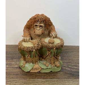 May include: A detailed figurine of an orangutan playing the drums. The orangutan has brown fur and is seated behind two drums. The base of the figurine has the name "Ringo" inscribed. The drums are beige and the base is green.