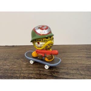 May include: A yellow textured character figure with a single tooth, wearing a green helmet with a red target symbol. The figure holds a red toy weapon and is positioned on a gray skateboard with white wheels, featuring a black grip tape surface.