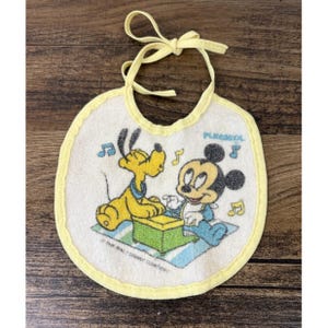 May include: Vintage baby bib featuring Mickey Mouse and Pluto playing music. The bib has a yellow trim and tie, with the text "PLAYSKOOL" and "THE WALT DISNEY COMPANY" printed on it. The bib is white with a colorful illustration.