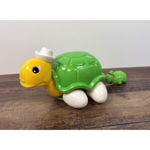 May include: A yellow and green toy turtle with a white hat and white feet. The turtle has a green shell and a smaller green turtle attached by a string. The toy is on a wooden surface.