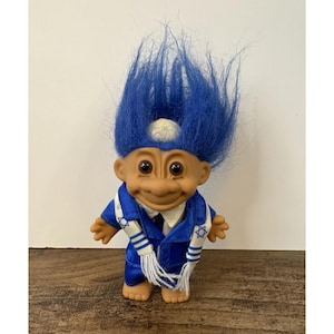 May include: A vintage troll doll with blue hair and a smiling expression. The doll is wearing a blue satin suit with a white and blue scarf featuring Star of David symbols. The troll has a white skullcap.