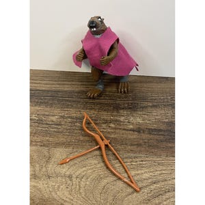May include: A brown anthropomorphic beaver action figure with a dark nose and visible teeth, wearing a magenta fabric cape. The figure has grey wrist bands and is accompanied by a separate brown plastic bow and arrow accessory.