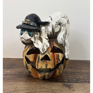 May include: A decorative Halloween figurine featuring a carved orange pumpkin base with a distressed finish. Perched on top is a white ghost wearing a black witch hat with a gold buckle, featuring a spider web and spider detail on its back.