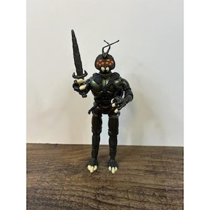May include: A black action figure of a humanoid insect warrior holding a black sword. The figure has a segmented body, insect-like head with multiple orange eyes, and white claws. The figure is posed on a wooden surface.