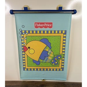 May include: A light blue Fisher-Price car window shade featuring a cartoon fish with yellow, blue, and green accents. The fish is inside a yellow and green frame. The shade has a blue top bar and suction cups.