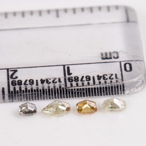 Puede incluir: Cuatro diamantes facetados de diferentes tama&ntilde;os y colores, incluyendo gris, amarillo y marr&oacute;n, se muestran sobre una superficie blanca junto a una regla marcada en cent&iacute;metros.
