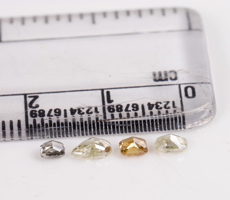 Puede incluir: Cuatro diamantes facetados de diferentes tama&ntilde;os y colores, incluyendo gris, amarillo y marr&oacute;n, se muestran sobre una superficie blanca junto a una regla marcada en cent&iacute;metros.
