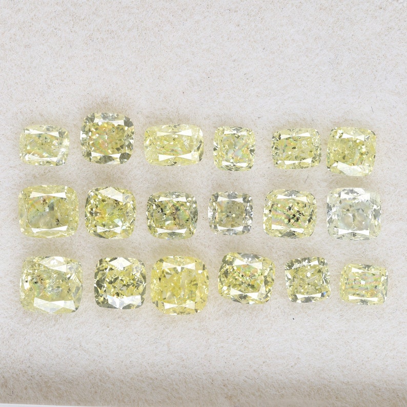 Op de afbeelding: Een verzameling van 16 kussen geslepen gele diamanten, gepresenteerd op een wit oppervlak. De diamanten zijn gerangschikt in vier rijen van vier.