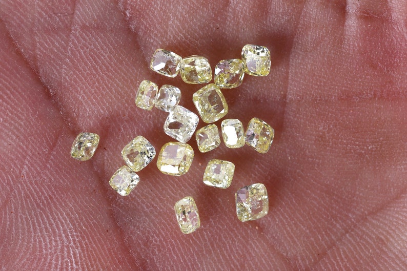 Op de afbeelding: Een verzameling kleine, gele, gefacetteerde edelstenen, waarschijnlijk diamanten, die op een hand worden getoond. De stenen zijn in kussenvorm geslepen.