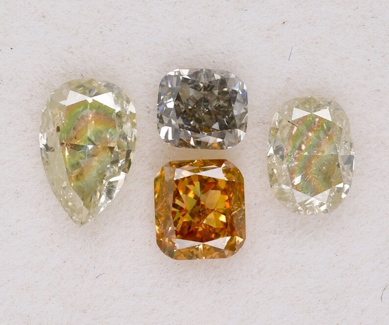 Puede incluir: Cuatro diamantes sueltos de diferentes formas y colores. Un diamante de forma pera, un diamante cuadrado de talla coj&iacute;n, un diamante redondo brillante y un diamante cuadrado de talla esmeralda. Los diamantes est&aacute;n todos sobre un fondo blanco.