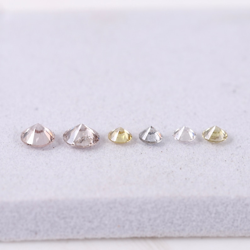 K&ouml;nnte beinhalten: Sechs runde, facettierte Diamanten in verschiedenen Gr&ouml;&szlig;en und Farben, darunter Braun, Gelb und Wei&szlig;, auf einer wei&szlig;en Oberfl&auml;che.