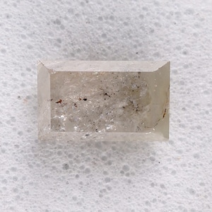 May include: A rectangular, light-colored diamond with a slightly cloudy appearance. The gemstone has a flat top and sides, with visible inclusions and imperfections. The diamond is set against a white background.