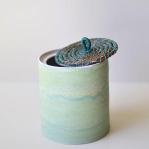 May include: A cylindrical ceramic container with a lid. The container is painted in shades of light green and blue. The lid is made of woven material in shades of brown and teal.