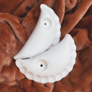 May include: Two white ceramic pieces shaped like crescent moons with a hole in the center. The pieces are on a brown fuzzy surface.