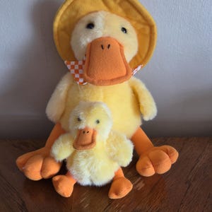 May include: A yellow plush duck with a yellow hat and a red and white checkered scarf, holding a smaller yellow plush duck.