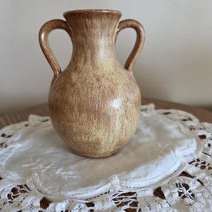 May include: A small, two-handled vase with a speckled brown and tan finish. The vase has a rounded body and a narrow neck. It sits on a white, lace doily with a floral pattern. The vase is likely ceramic or pottery.