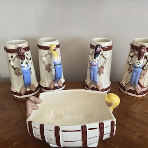 May include: A set of ceramic barware featuring four beer stein-shaped mugs and a rectangular serving bowl. The mugs have a cream base with a brown rim and a painted figure. The bowl is white with brown accents and yellow handles.