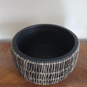 May include: A round decorative bowl with a black interior and a woven exterior. The exterior is made of interwoven light brown and silver-colored metal wires. The bowl sits on a wooden surface.
