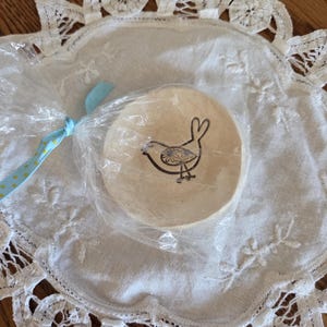 May include: A round, beige coaster with a black bird design, packaged in clear plastic and tied with a blue ribbon. The coaster sits on a white, lace-edged doily.