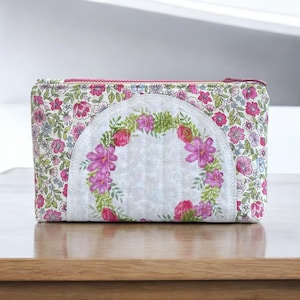Puede incluir: Una bolsa blanca con cremallera con estampado floral y diseño acolchado. La bolsa presenta una corona floral circular en el centro.