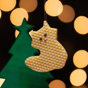 Może przedstawiać: Zielona ozdoba choinkowa i żółta ozdoba w kształcie skarpety o wzorze plastra miodu wiszą na rozmytym tle ciepłych, świecących świateł. Skarpeta ma dwa małe, ciemne zdobienia. Ozdoby znajdują się na drewnianej powierzchni.