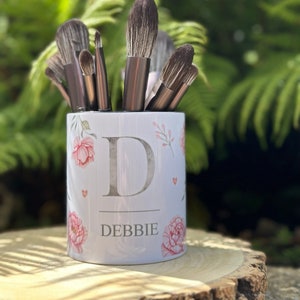 May include: A white ceramic makeup brush holder with a floral design and the name "DEBBIE" printed on it. The holder is filled with a variety of makeup brushes.