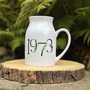 May include: White ceramic mug with a black 1973 design featuring green foliage accents. The mug has a handle and is resting on a wooden surface.