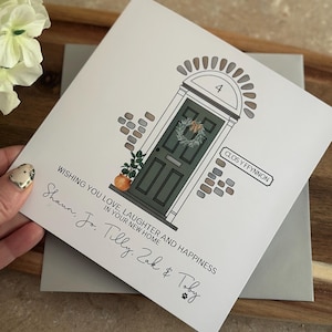 May include: A white card with a hand-drawn illustration of a green door with a wreath. The door has a number "4" above it and the text "CLOS Y FFYNNON" on the right side. The card has the text "WISHING YOU LOVE, LAUGHTER AND HAPPINESS IN YOUR NEW HOME" and the names "Shaun, Jo. Tilly. Zak & Toby" written in cursive.