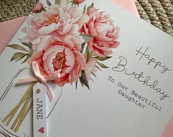 May include: A white birthday card with a watercolor illustration of pink peonies in a mason jar. The card reads "Happy Birthday To Our Beautiful Daughter." A small tag with the name "JANE" is attached with a pink ribbon.