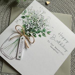 May include: A square greeting card featuring a watercolor illustration of a bouquet of white baby's breath and green eucalyptus in a glass jar. A jute twine bow holds a white tag with the name JESSICA and a heart. Text reads Happy Birthday To Our Beautiful Daughter.