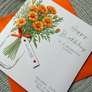 May include: A white birthday card featuring a bouquet of orange marigold flowers in a glass jar illustration. An orange ribbon ties a white tag with the name ELAINE and a red heart. Text reads Happy Birthday To A Wonderful Sister and October 2026 Birth Flower Marigold.
