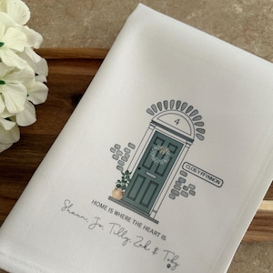 May include: White tea towel featuring an illustration of a green door. The door has a wreath and a sign that reads "CLOS Y FFYNNON". The text "HOME IS WHERE THE HEART IS" is printed below the door. The names "Shaun, Jo, Tilly, Zak & Toby" are handwritten below the text.