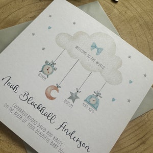 May include: A white card with blue and gray watercolor illustrations of a cloud, a moon, a star, and a clock. The text on the card reads "Welcome to the world", "Noah Blackhall Anderson", "Congratulations David and Kirsty on the birth of your beautiful baby boy", "8:29 PM", "12.05.24", and "4LBS 10ZS".