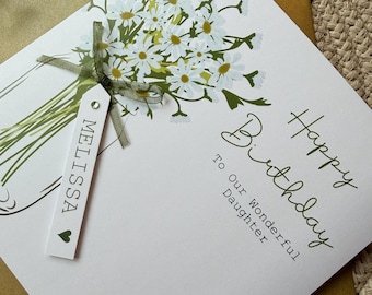 May include: A white birthday card featuring a bouquet of white daisies in a glass jar illustration, a green ribbon, and a white tag with the name MELISSA and a heart icon. The text reads Happy Birthday To Our Wonderful Daughter.