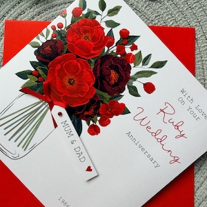 May include: A white Ruby Wedding Anniversary card featuring a floral design of red roses and greenery. The card includes the text "With Love On Your Ruby Wedding Anniversary", "Mum & Dad", and the years "1986 - 2026".