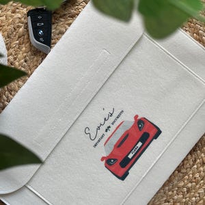 May include: Cream-colored document holder with a red sports car graphic and the name "Flora" on the license plate. The holder is open, revealing a stitched seam and the text "Evie's Important Documents."