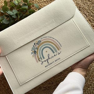 May include: A cream-coloured maternity notes holder with a flap. It features a rainbow design with floral accents and the name "Sophie's" in script, along with "Maternity Notes". The text "Our journey to meet baby Clayton-Priestley" is also visible.