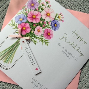May include: A white greeting card featuring a watercolor illustration of pink, purple, and blue cosmos flowers in a glass jar. A pink ribbon ties a white tag labeled CHARLOTTE with a heart. Text reads Happy Birthday To A Wonderful Friend October 2026 Birth Flower Cosmos.