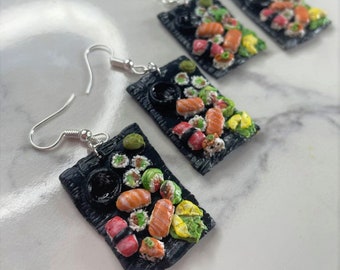 May include: Sushi-themed earrings with miniature sushi pieces on black rectangular plates. The earrings display various sushi types, including nigiri and rolls, with vibrant colours and detailed craftsmanship. The earrings have silver-coloured fish hook closures.