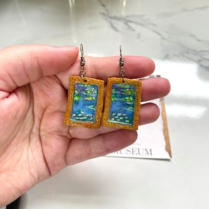 以下が含まれることがあります： 青い池に睡蓮が描かれた絵がついた、金色のフレームのイヤリングのペア。イヤリングはブロンズのフックからぶら下がっています。