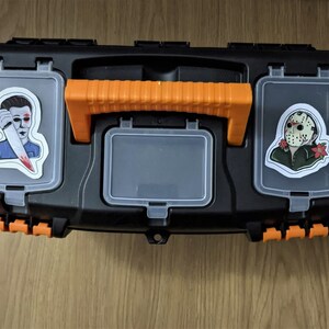 K&ouml;nnte beinhalten: Eine schwarze Werkzeugkiste mit orangefarbenen Griffen und zwei Aufklebern. Ein Aufkleber zeigt eine Figur mit einem blutigen Messer und der andere Aufkleber zeigt eine Figur mit einer Hockeymaske.