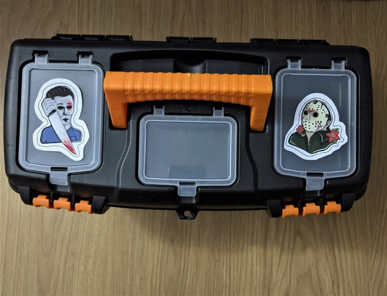 K&ouml;nnte beinhalten: Eine schwarze Werkzeugkiste mit orangefarbenen Griffen und zwei Aufklebern. Ein Aufkleber zeigt eine Figur mit einem blutigen Messer und der andere Aufkleber zeigt eine Figur mit einer Hockeymaske.