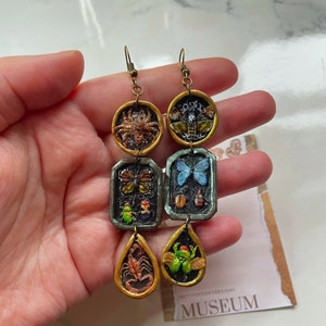 Puede incluir: Un par de aretes largos y colgantes con un esquema de color dorado y verde. Cada arete presenta tres dijes: un dije redondo con un insecto pintado, un dije rectangular con una mariposa pintada y otros insectos, y un dije en forma de lágrima con un escorpión o una rana pintados. Los aretes están etiquetados como "PRETTYSPOOKYDESIGNS MUSEUM".