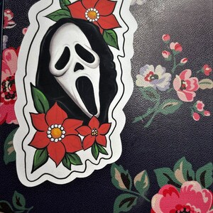 K&ouml;nnte beinhalten: Ein wei&szlig;er Aufkleber mit einer Schwarzwei&szlig;illustration eines Geistes, der eine Maske mit einer roten Blume oben tr&auml;gt. Der Aufkleber befindet sich auf einem schwarzen Hintergrund mit einem Blumenmuster.