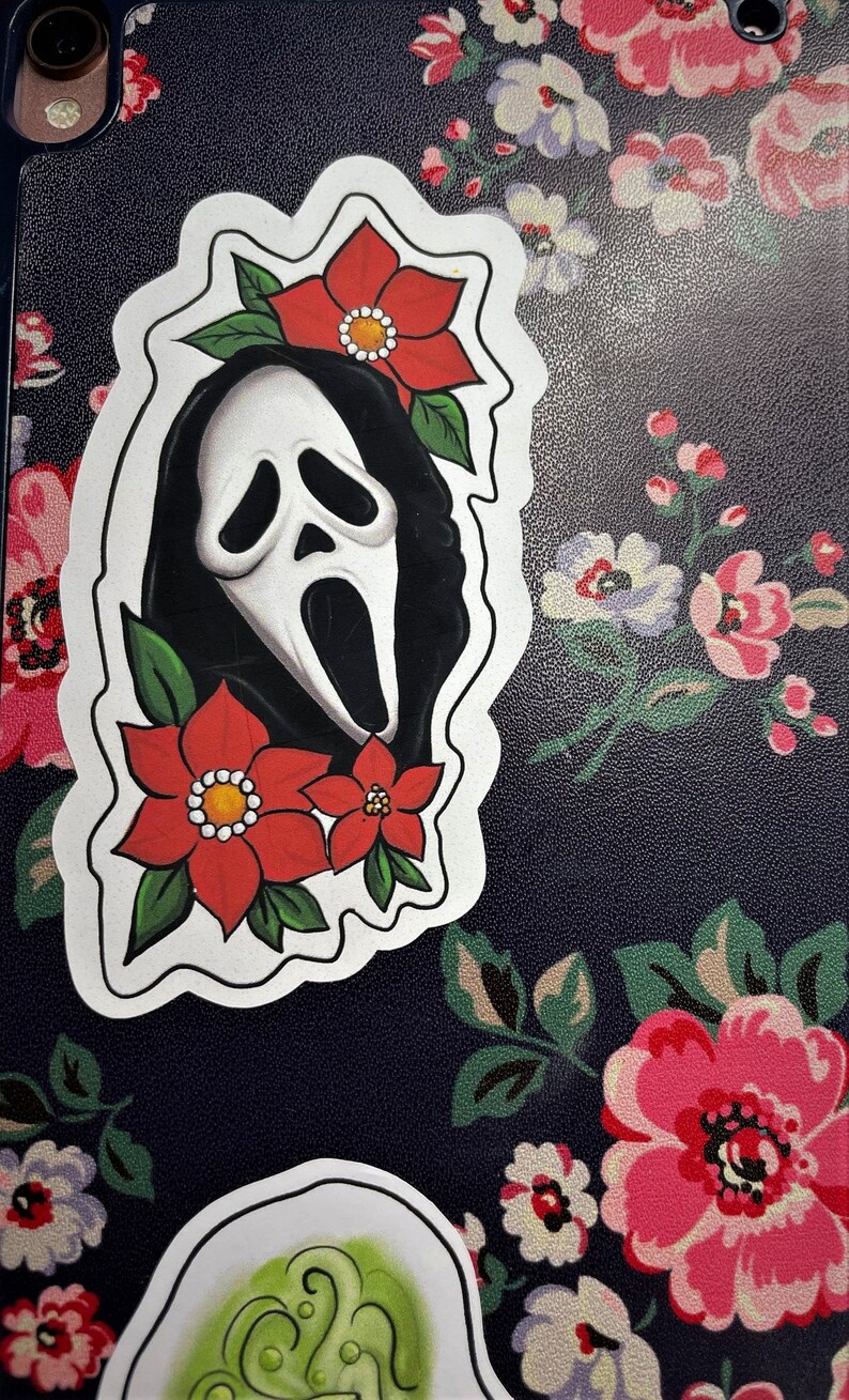 K&ouml;nnte beinhalten: Ein wei&szlig;er Aufkleber mit einer Schwarzwei&szlig;illustration eines Geistes, der eine Maske mit einer roten Blume oben tr&auml;gt. Der Aufkleber befindet sich auf einem schwarzen Hintergrund mit einem Blumenmuster.