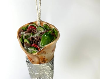 May include: A handmade ornament shaped like a wrapped kebab, with a brown pita, meat, lettuce, tomato, cucumber, and a green pepper. The ornament is wrapped in silver foil and hangs from a white string.