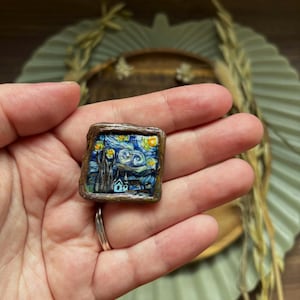 May include: A square ring featuring a miniature painting of Van Gogh's "Starry Night." The artwork is set within a brown, textured frame. The ring's band is silver-colored.