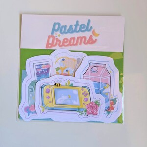 May include: A set of four pastel-colored stickers featuring a video game console, a milk carton, a night sky scene, and a floral design. The stickers are on a white background with the text "Pastel Dreams" in blue and pink.