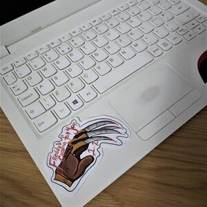 K&ouml;nnte beinhalten: Ein wei&szlig;er Laptop mit einem Freddy Krueger-Aufkleber auf der rechten Seite des Touchpads. Der Aufkleber zeigt eine Zeichnung von Freddys Handschuh mit den Worten "Freddy's is coming for you" und "7-2" darauf.